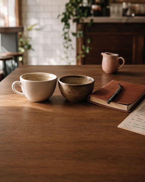 Two coffee cups, a leather notebook with a pen, and a handwritten task list on a warm wooden cafe table.